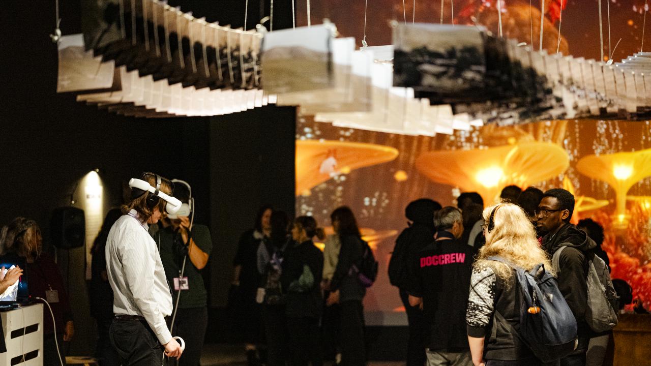 A dimly lit exhibition room with a large orange screen on the back wall. A crowd stand around wearing VR headsets.