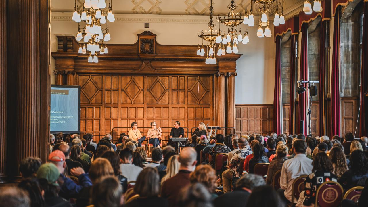 Busy crowd of people facing away from the camera look towards 4 people sat on chairs at the front of a large, grand hall.
