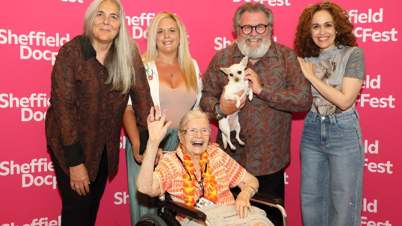 Five people are shown in front of a pink backdrop with the Sheffield DocFest logo repeated on it in white. One of the people is sat in a wheelchair with their hand up in a wave and another person, stood behind holds a white chihuahua. 