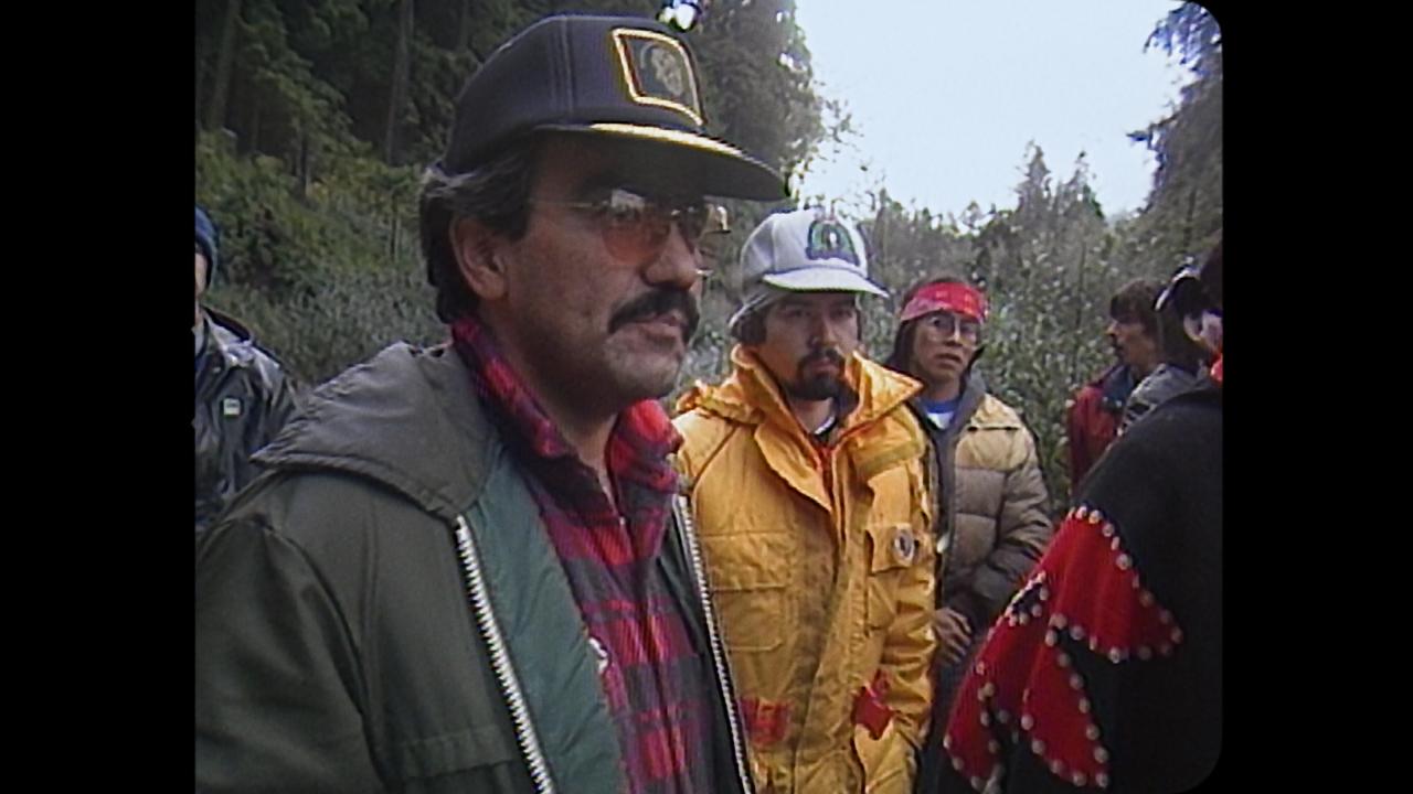 Still from The Stand - three people stand in a diagonal line wearing heavy outdoor clothing including caps, big coats and checked shirts. Behind them is a woodland of thick green trees.