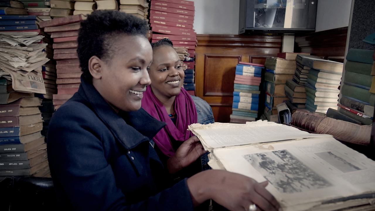 A small room with books stacked high. Two women in coats and scarves stand in the middle of the frame with large smiles on their face. The woman closest to the camera holds a large old book open and is looking down at it.