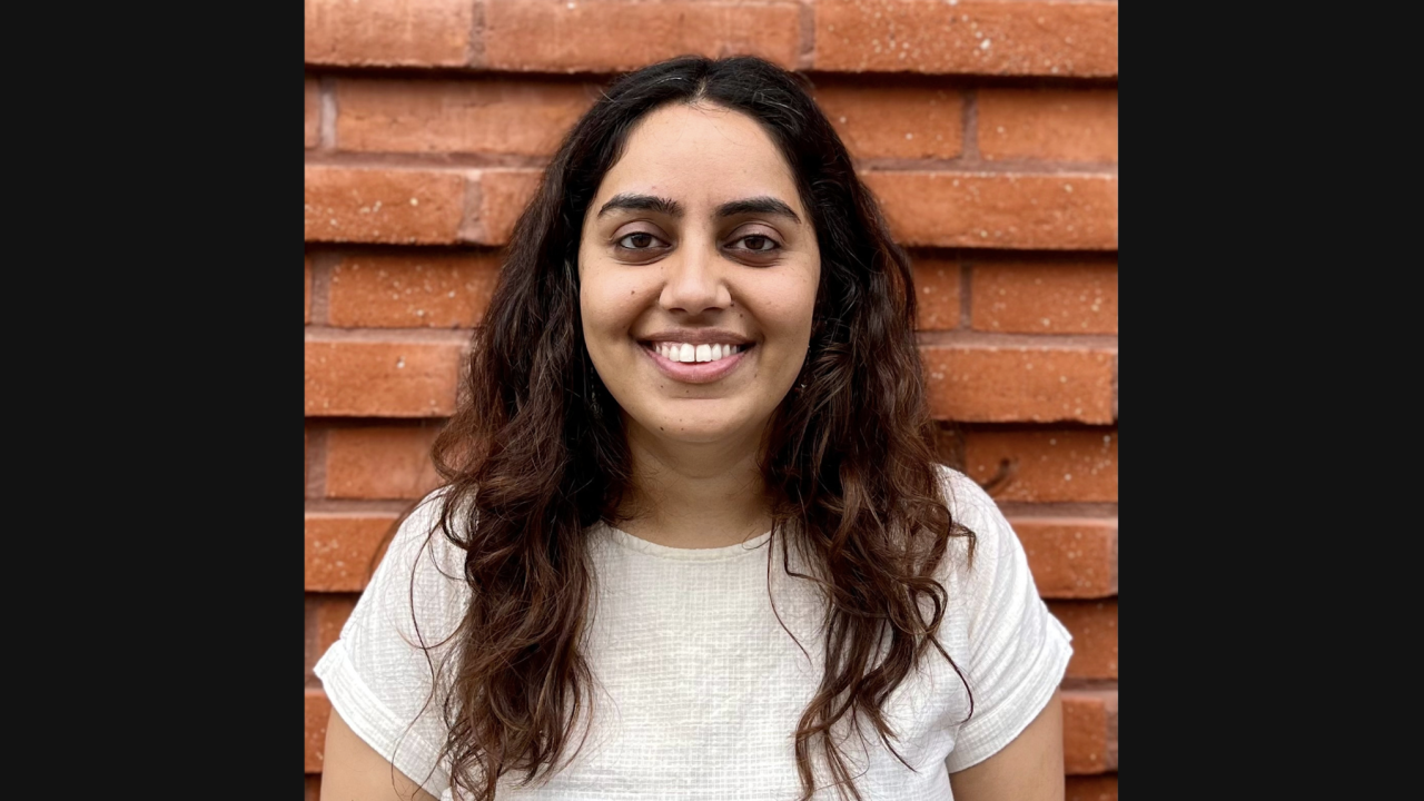 Mita Suri headshot. Image of a woman with dark curly hair. She's wearing a white top and is pictured against a brick wall.