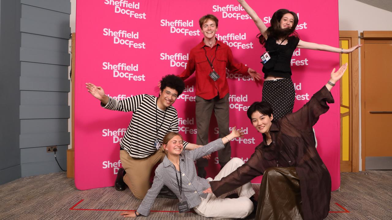 5 young people jumping in the air in front of a pink step and repeat with the Sheffield DocFest logo on.