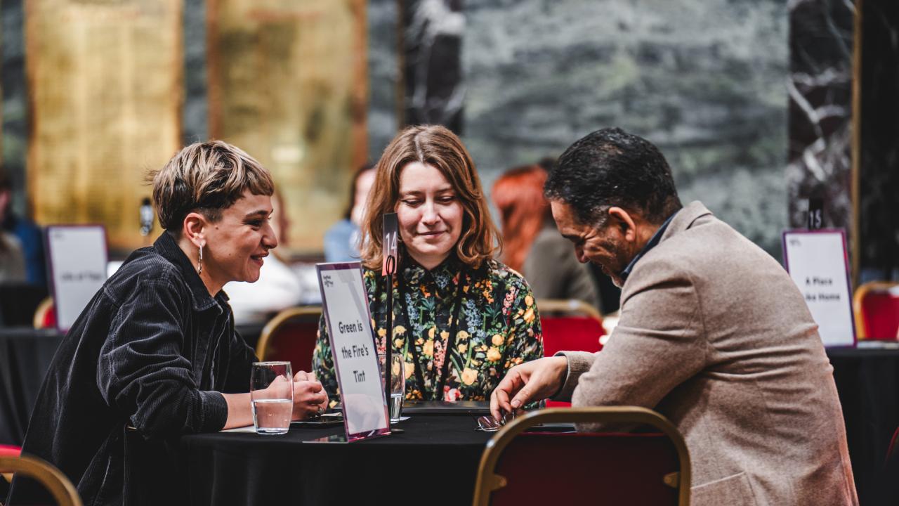 Three people are sat around a table, one is facing towards the camera and is wearing a floral shirt. There’s a woman with short hair and a black jacket smiling at a man across the table, who’s wearing a camel jacket and is playing with a pair of glasses on the table. 