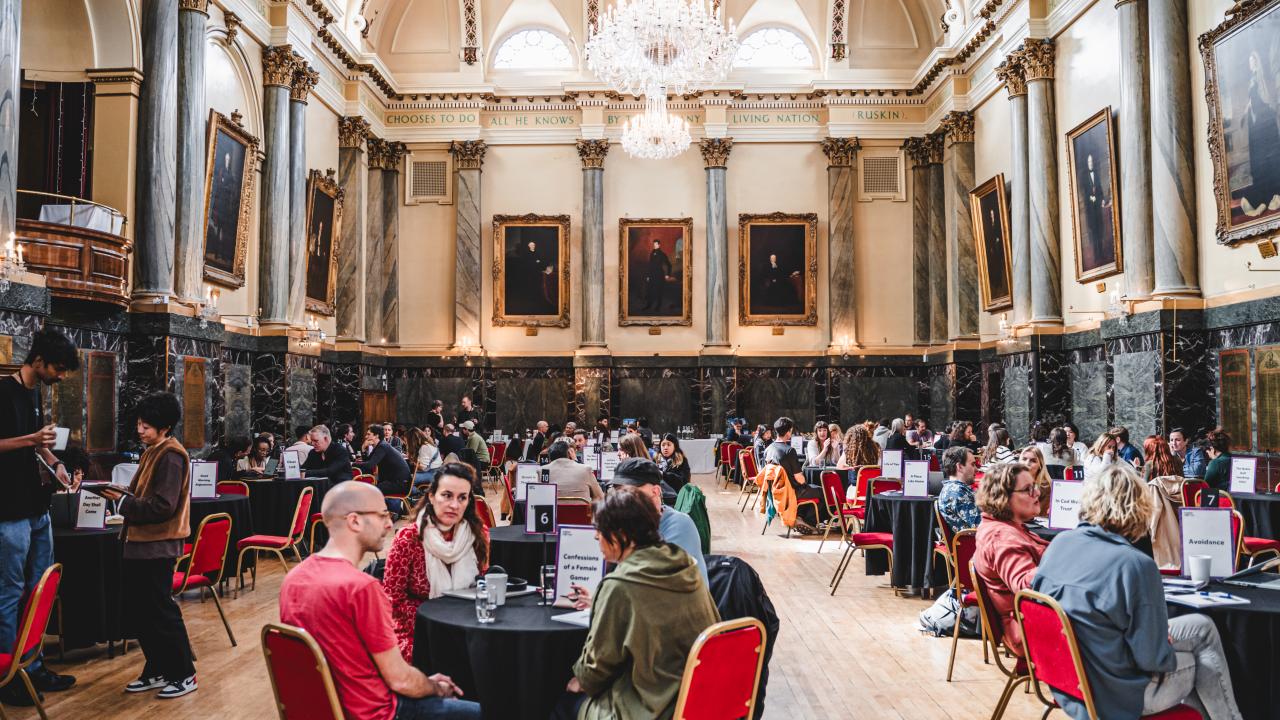 A busy grand hall full of people talking around tables sat on red velvet chairs.