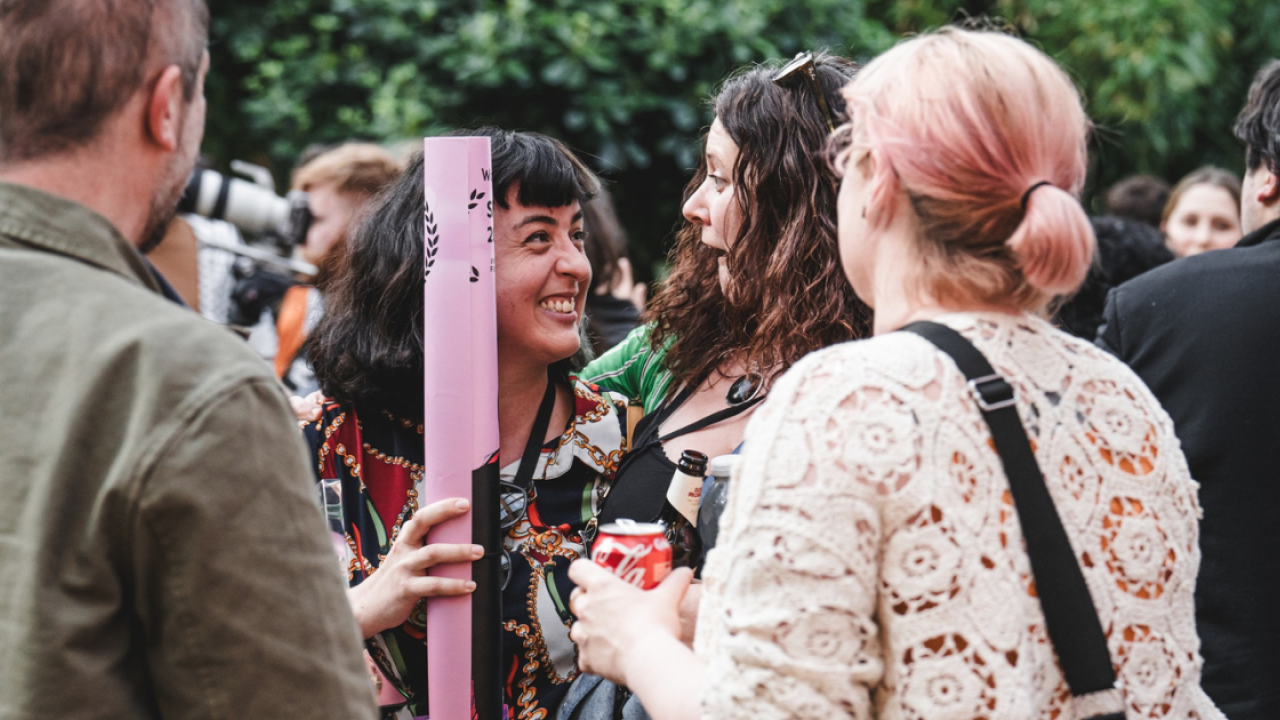 People surrounded by greenery at a busy drinks reception. In the centre of the photo two people looking at each other smile widely. The person on the left is holding a film poster rolled up in their hand.