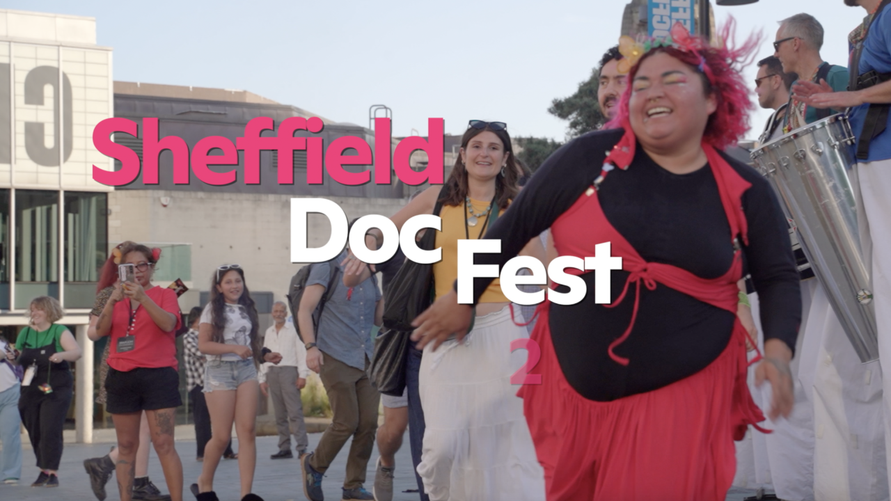 A busy town square with people laughing and dancing. On the far right of the image two men on stilts are banding drums.