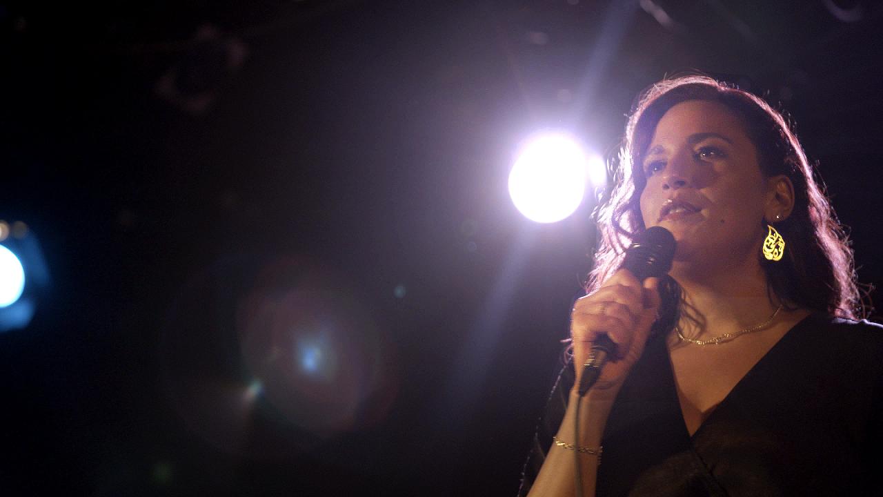 On the right hand side of a frame, a woman holding a microphone stands on a stage with a spotlight on her.