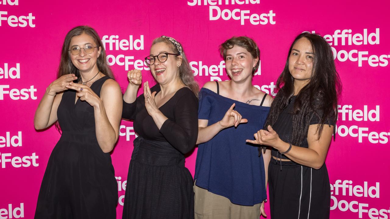 Four people stand in front of a in front of a pink step and repeat with the Sheffield DocFest logo on.