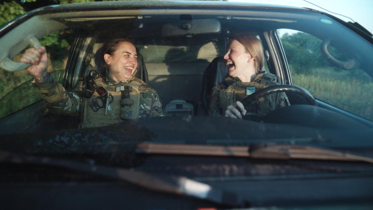 Two women wearing camouflaged clothing sit in the front seat of a car. The two women look at each other with wide smiles on their face. 