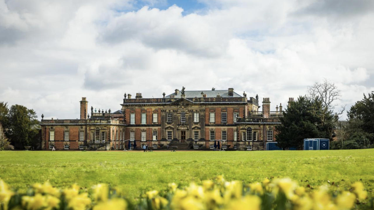 In the foreground, yellow flowers and a grassy lawn. In the distance a large country house sits against a blue sky filled with white clouds.