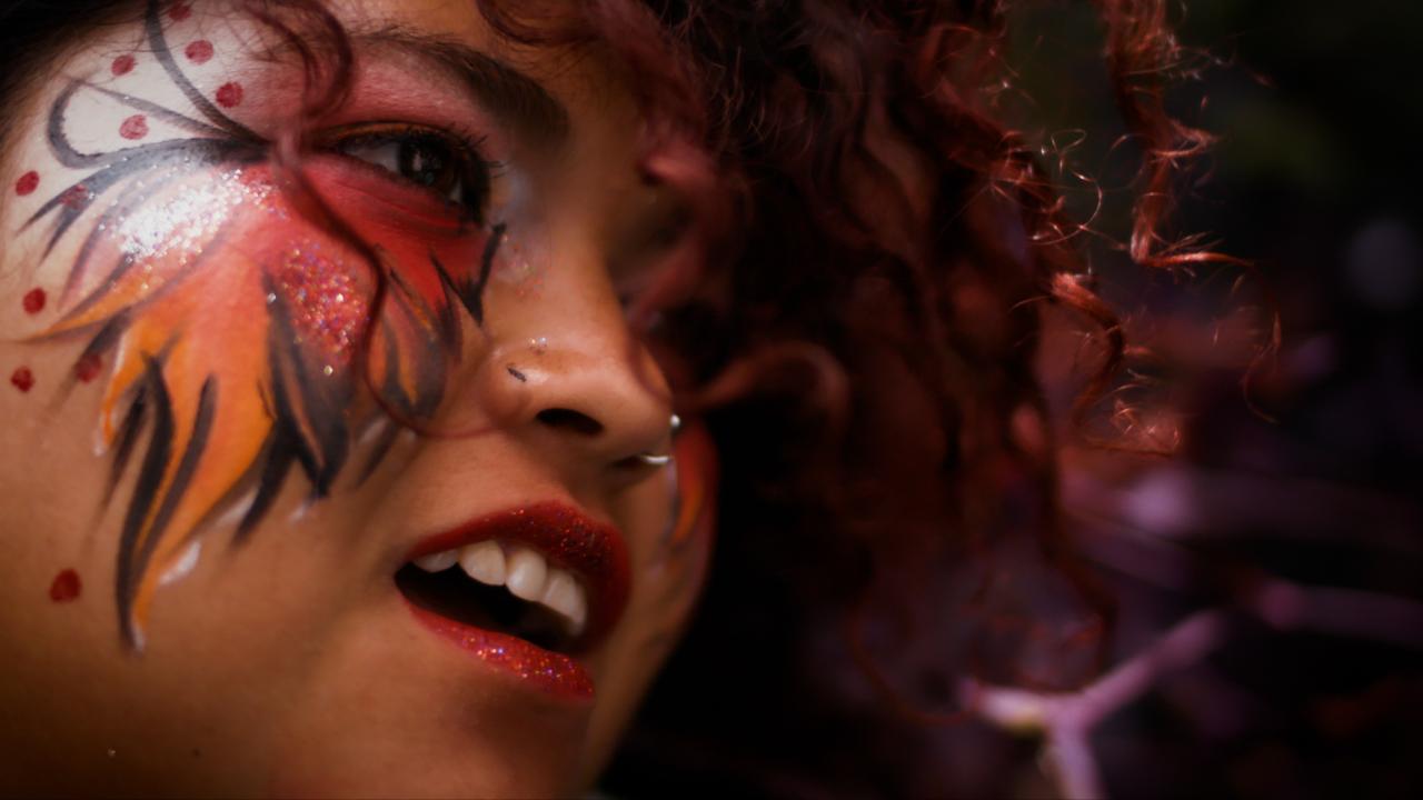 A close up photograph of a young girl with colourful face paint around her eyes, pink sparkly lipstick and a nose ring