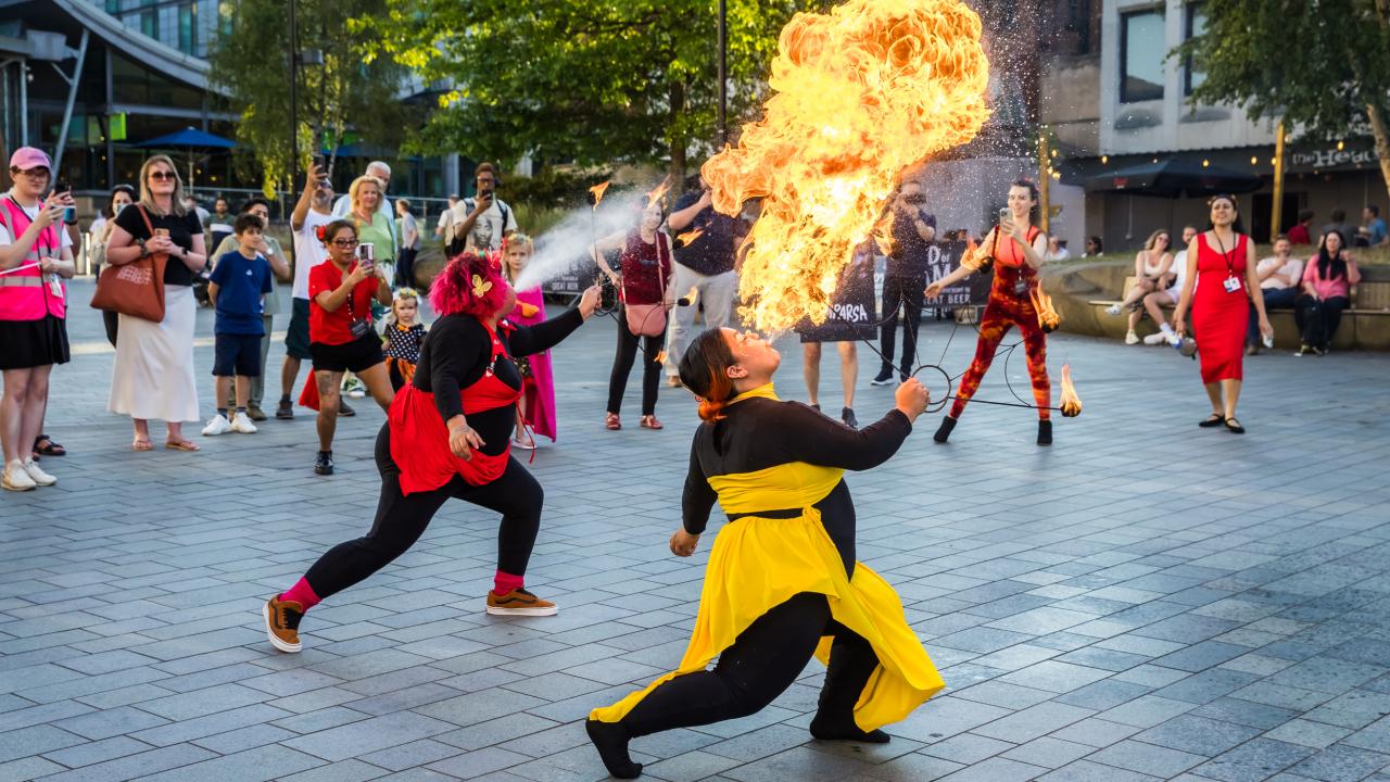 Two people breathe fire in a square whilst a crowd watches.