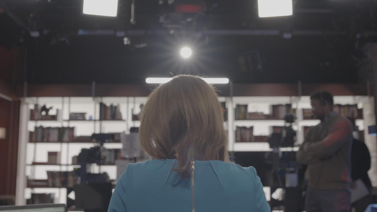 Viewed from behind, a woman with blonde hair in a blue top sits in a brightly lit television studio facing a bookshelf backdrop and production crew under professional set lights.