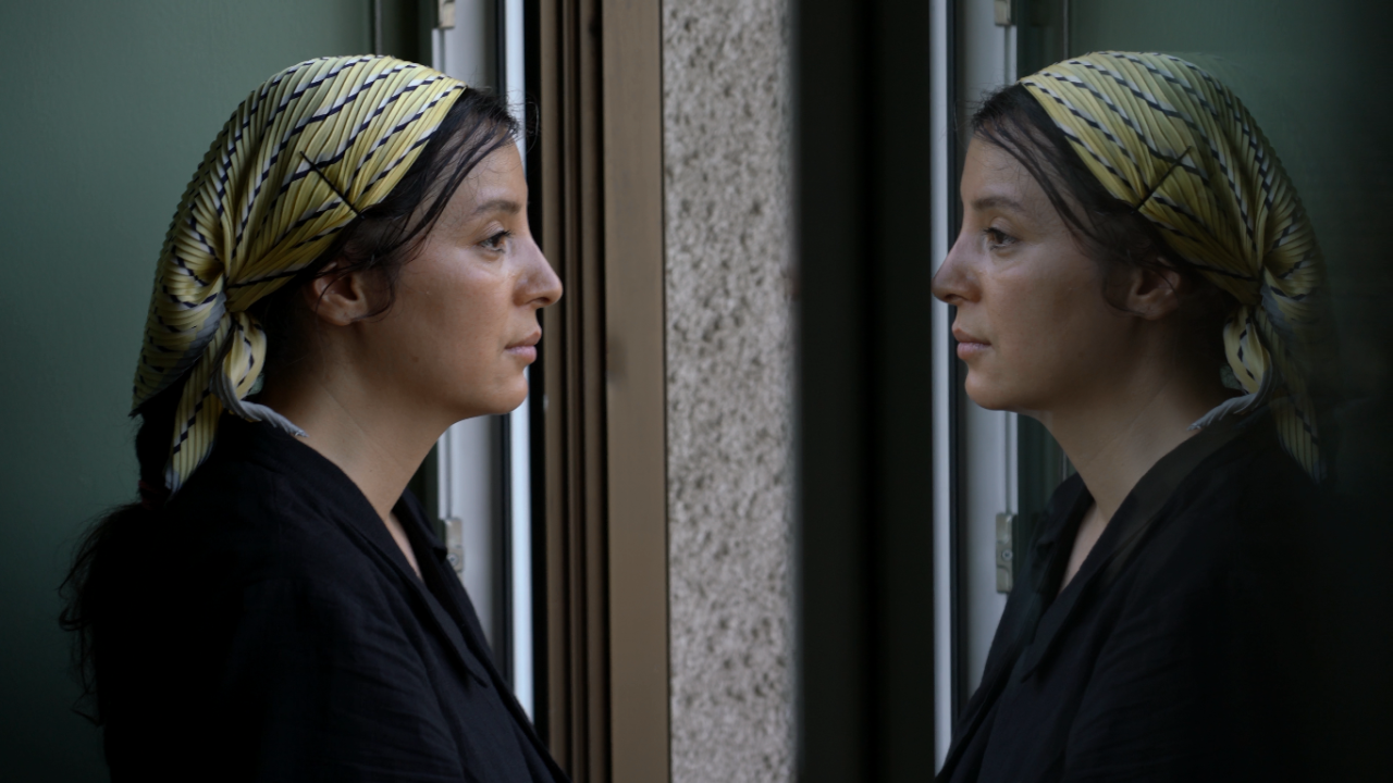 A woman in a yellow headscarft and black top stands on the left and looks at her reflection in a window.