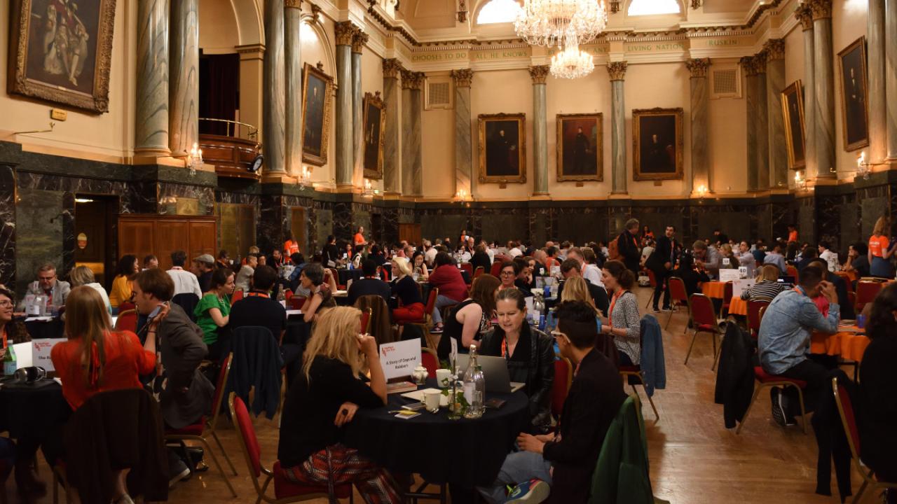 A crowded room full of people taking meetings around round tables