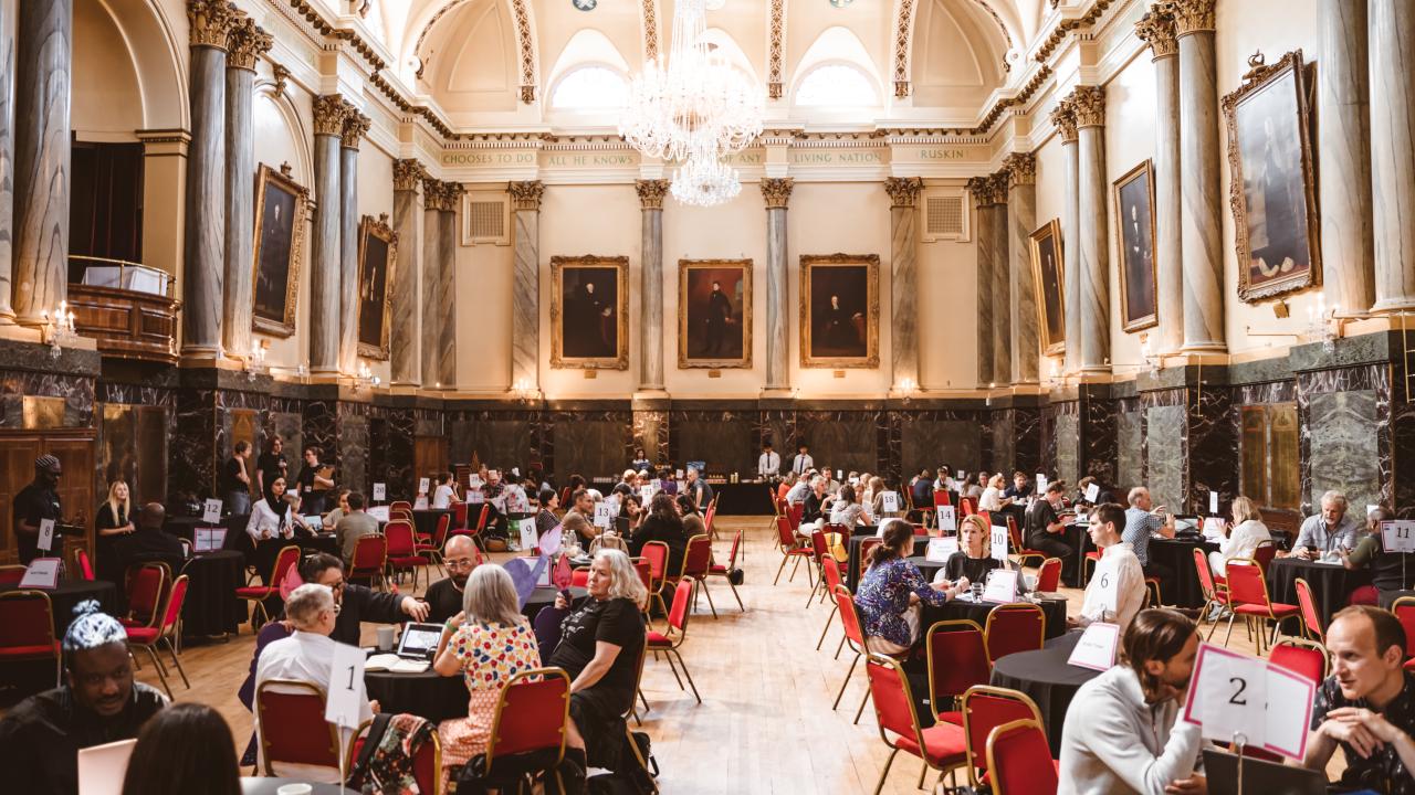 A crowded room full of people taking meetings around round tables