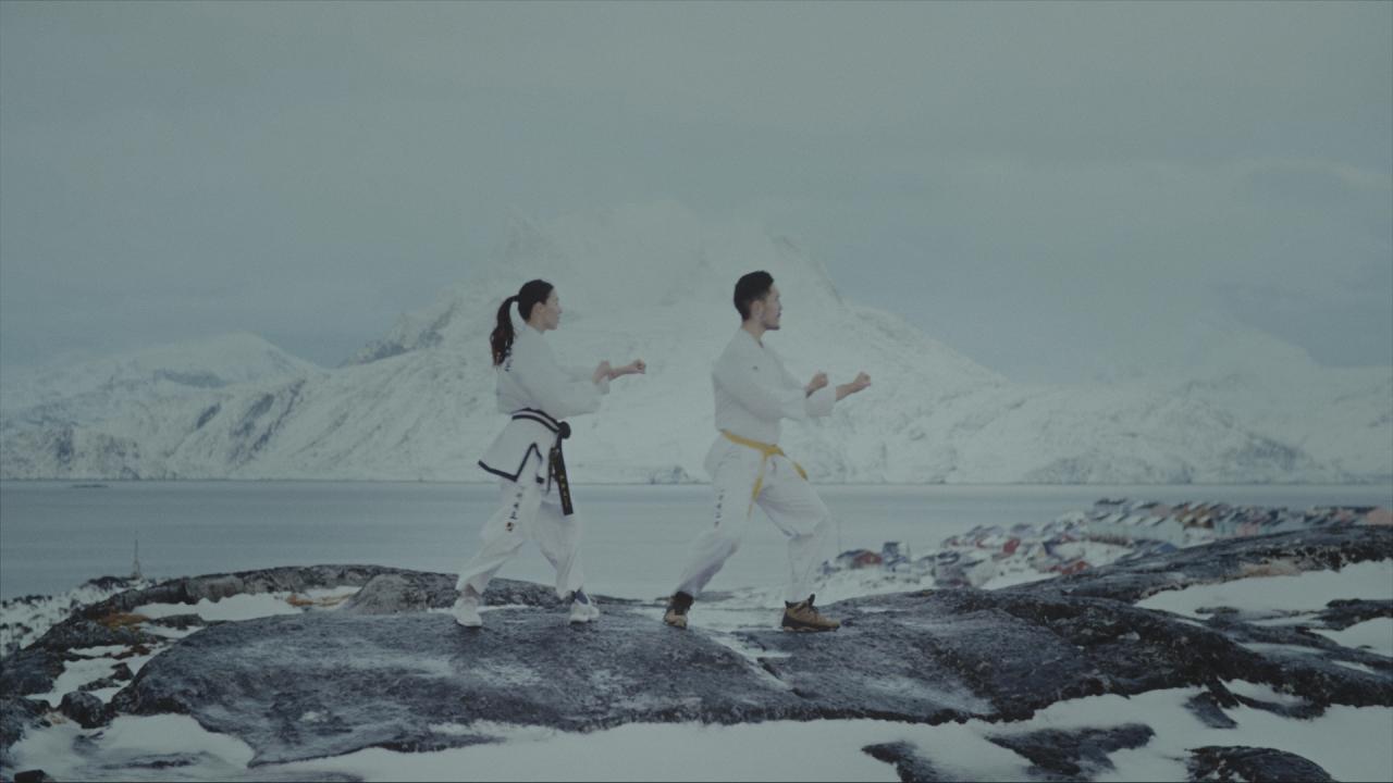 Two people in white martial arts uniforms practice synchronized movements on a rocky, snow-covered landscape, with a calm body of water and snow-capped mountains in the background.