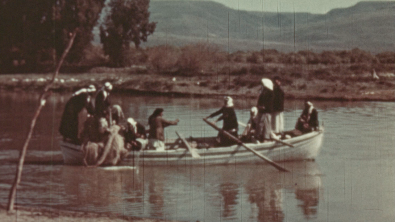 A group of people and animals in small boats crossing a calm river, with a rural landscape and hills in the background.