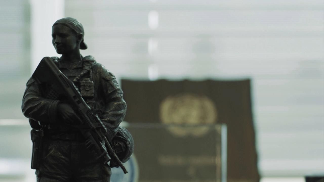 A small statue of a female soldier holding a rifle, with a blurred podium and emblem in the background, suggesting an official or institutional setting.
