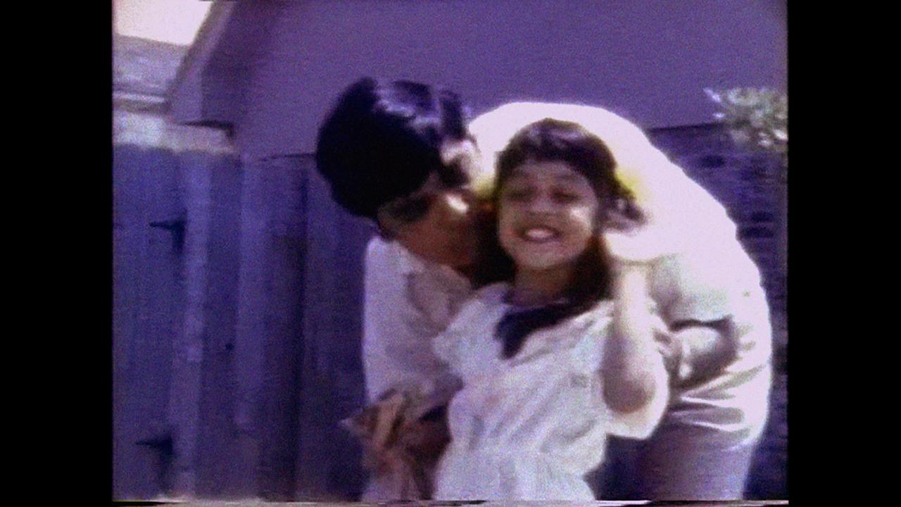 A still from The Gas Station Attendant: a home video image of a father kissing his young daughter on the cheek as she smiles. The image has the soft, faded quality of vintage camcorder footage.
