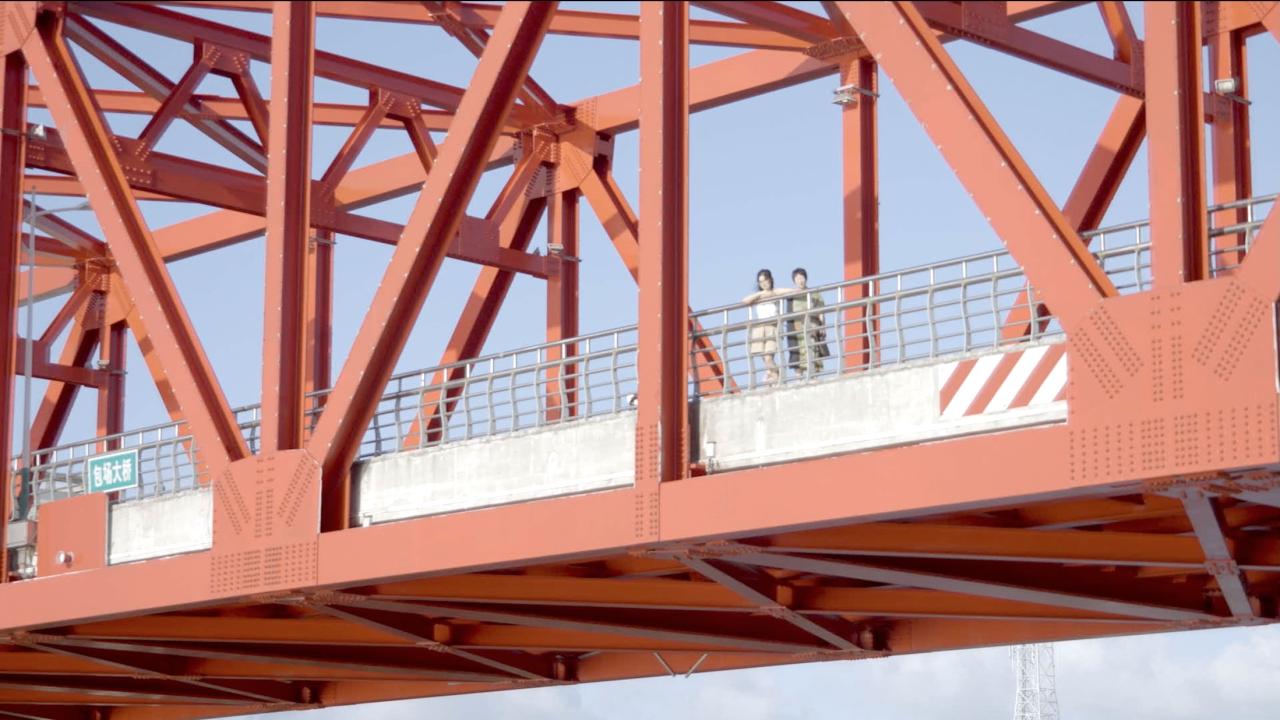 Two people stand side by side on a pedestrian walkway of a large red steel bridge, framed by its geometric beams, looking outward.