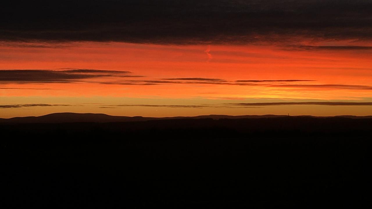 A bright orange-red sunset over dark, silhouetted hills with streaks of clouds above.