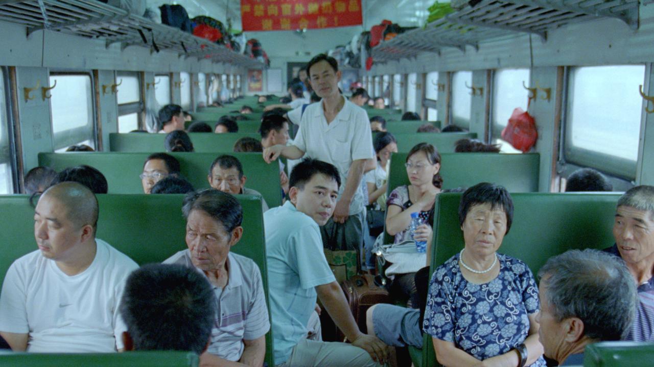 A crowded train carriage with rows of green seats filled with passengers. A man stands in the aisle while others sit facing each other, some looking toward the camera. 