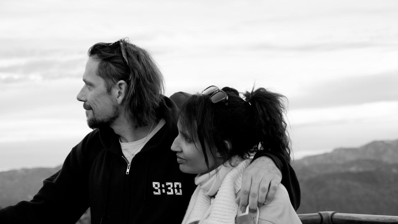 A couple stands close together at a scenic viewpoint, the man with his arm around the woman as they both look off into the distance, mountains stretching out behind them in a calm black and white setting.