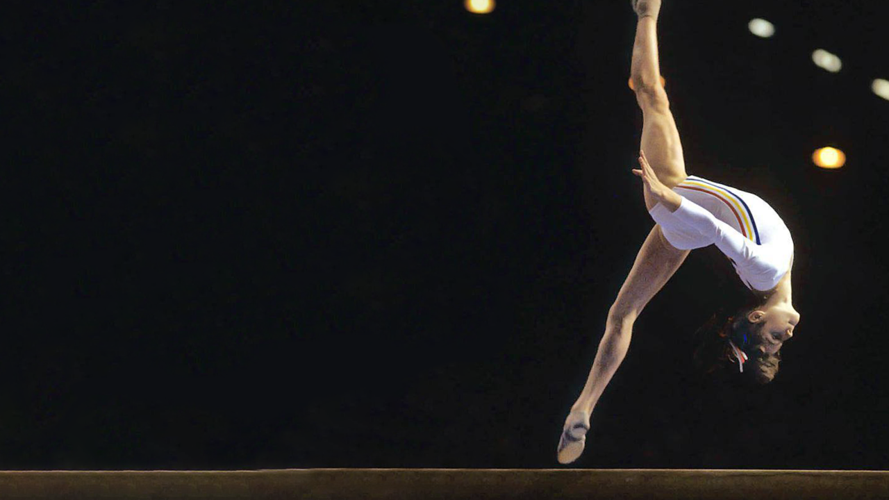 A gymnast in a white leotard performs a dynamic, arched split-leap mid-air above a balance beam against a dark, spotlighted arena background.