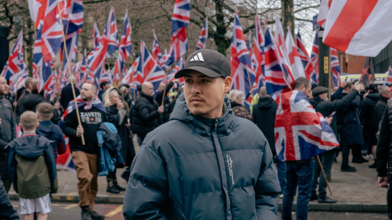 Anywhere but here  A man in a dark puffer jacket and cap stands in the foreground, looking to the side, while a large crowd behind him holds numerous Union Jack flags at a street protest.