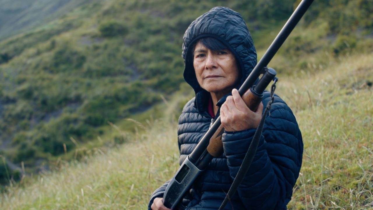 A person in a black puffer jacket holding a rifle stands in the middle of the frame. In the background are grassy hillsides.
