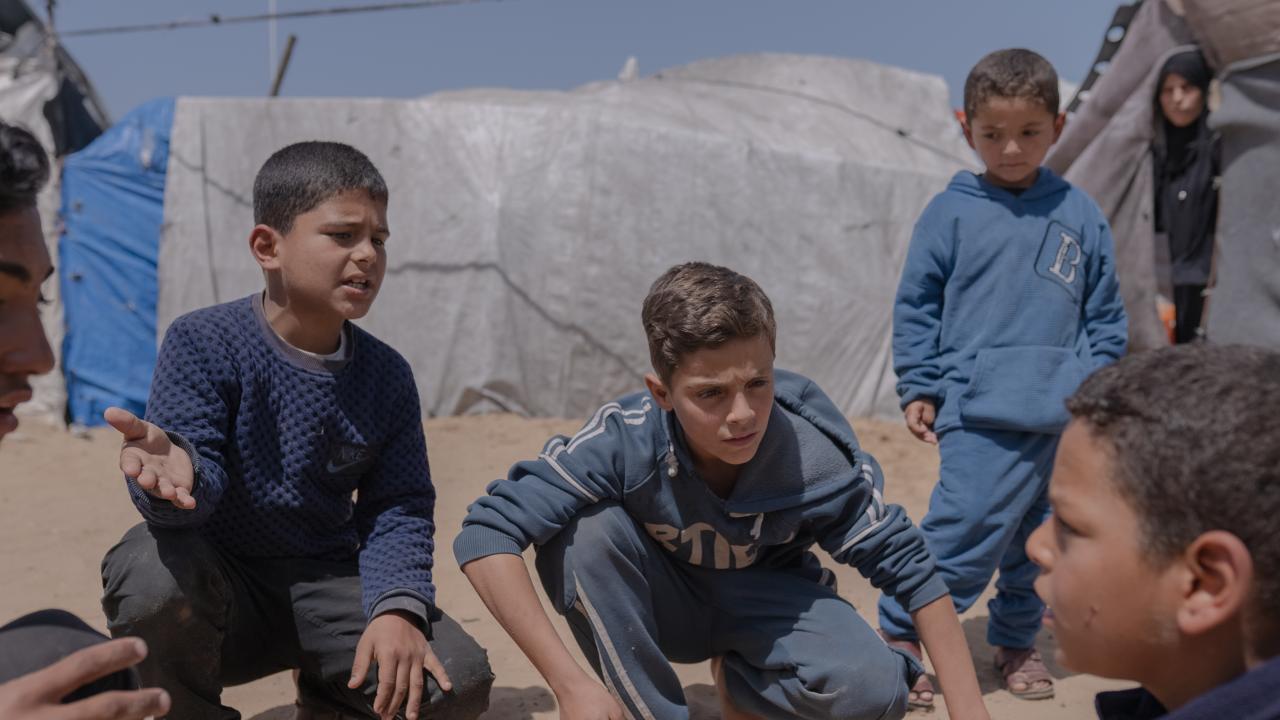 Several children gather and crouch in a sandy area between makeshift tents, appearing focused on a game or conversation. The scene takes place in a camp setting under a clear blue sky, with tarps and temporary shelters in the background.