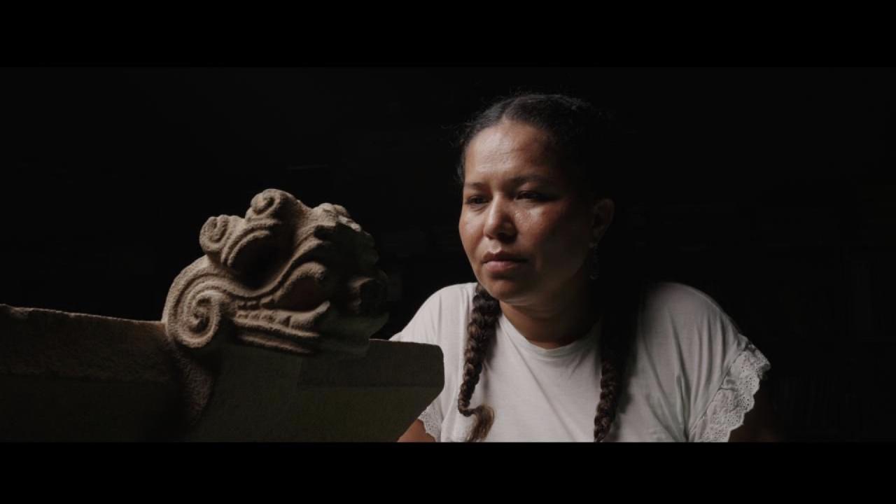 A woman with braided hair, wearing a light shirt, looks intently at a carved stone sculpture with a stylized face.