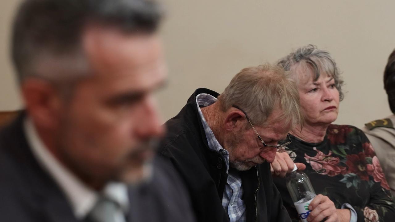 Three people seated indoors; a blurred man in front, while an older man looks down holding a bottle and an older woman looks ahead seriously.