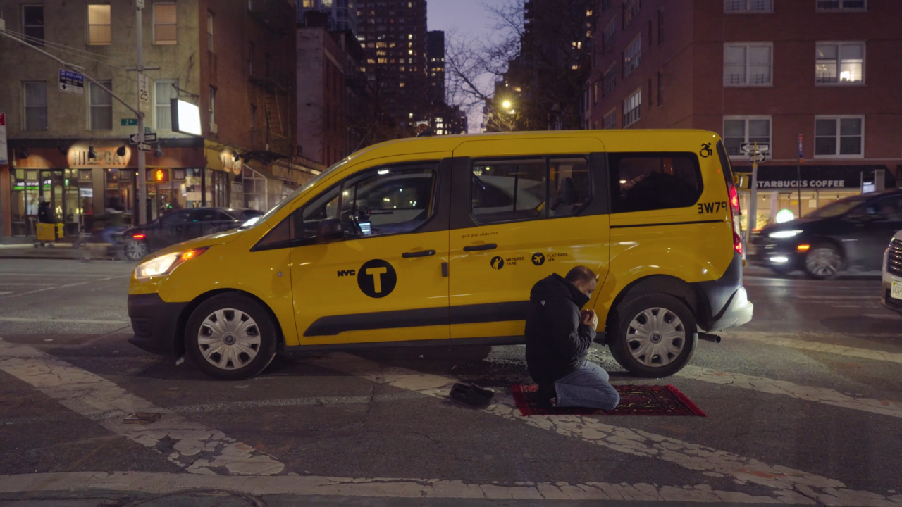 A yellow taxi minivan is parked centrally on a busy city street at dusk, with a man infront of it, praying on a prayer mat.