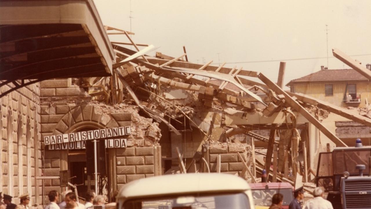 A partially collapsed building with debris and broken beams around a bar-restaurant entrance, while people are gathered nearby looking at the damage