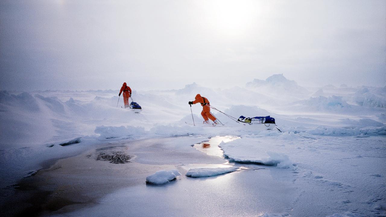 Two people in bright orange cold-weather suits ski across a vast, icy landscape in blowing snow, each pulling a sled behind them. 