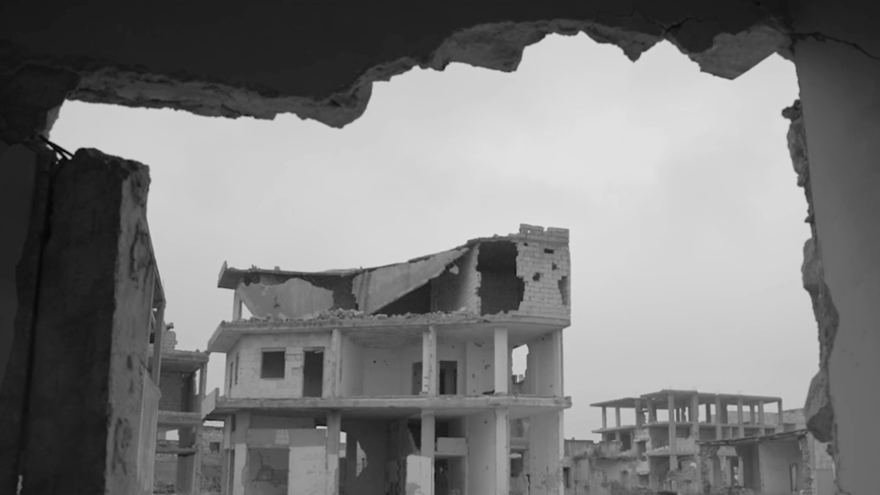 Black-and-white view of a heavily damaged building seen through a large hole in a cracked wall, with ruined structures in the background.