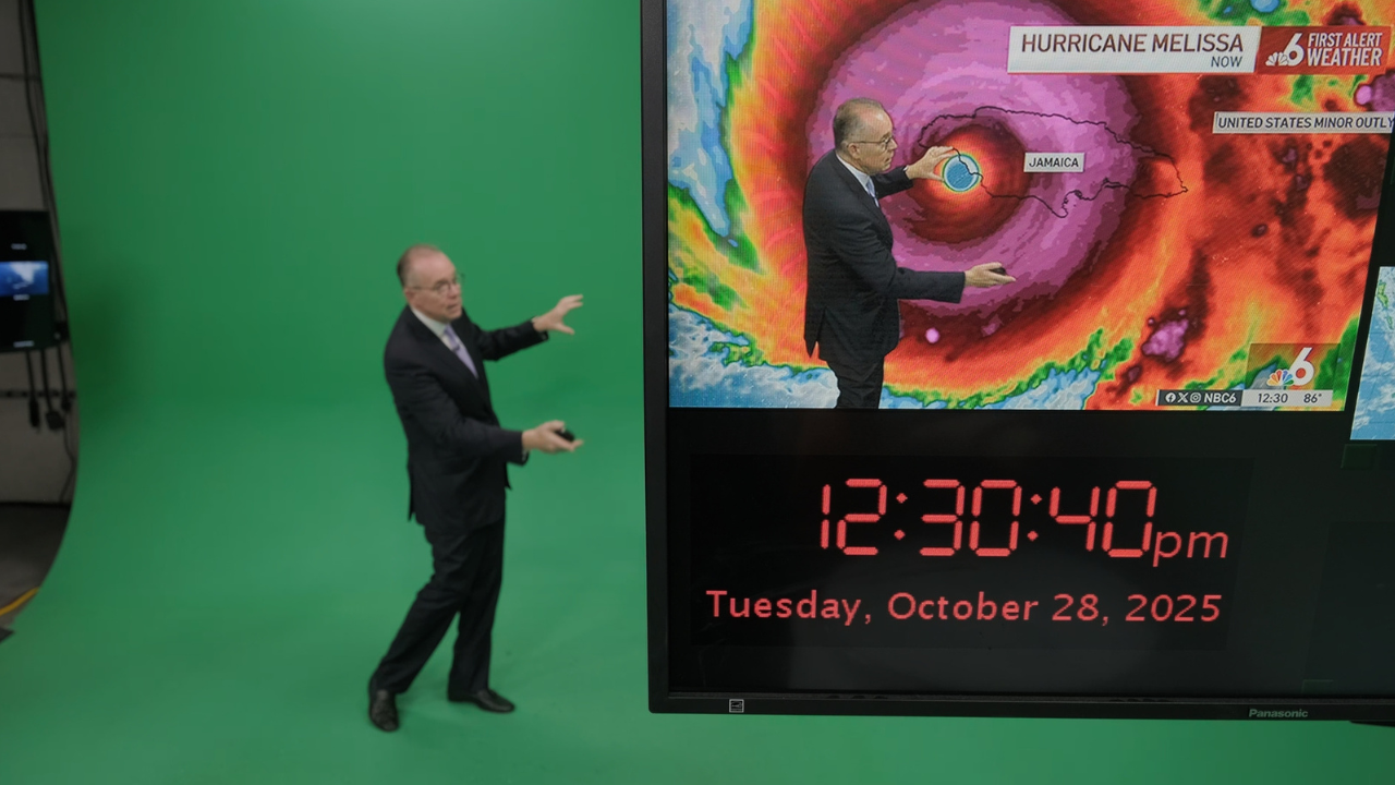 A weather presenter stands before a green screen, pointing at a monitor showing a hurricane over Jamaica.