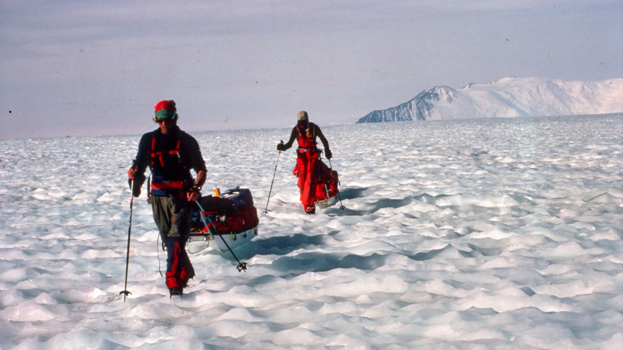 Two people in expedition clothes and walking poles trekking through deep snow, with snow mountains in the distance.