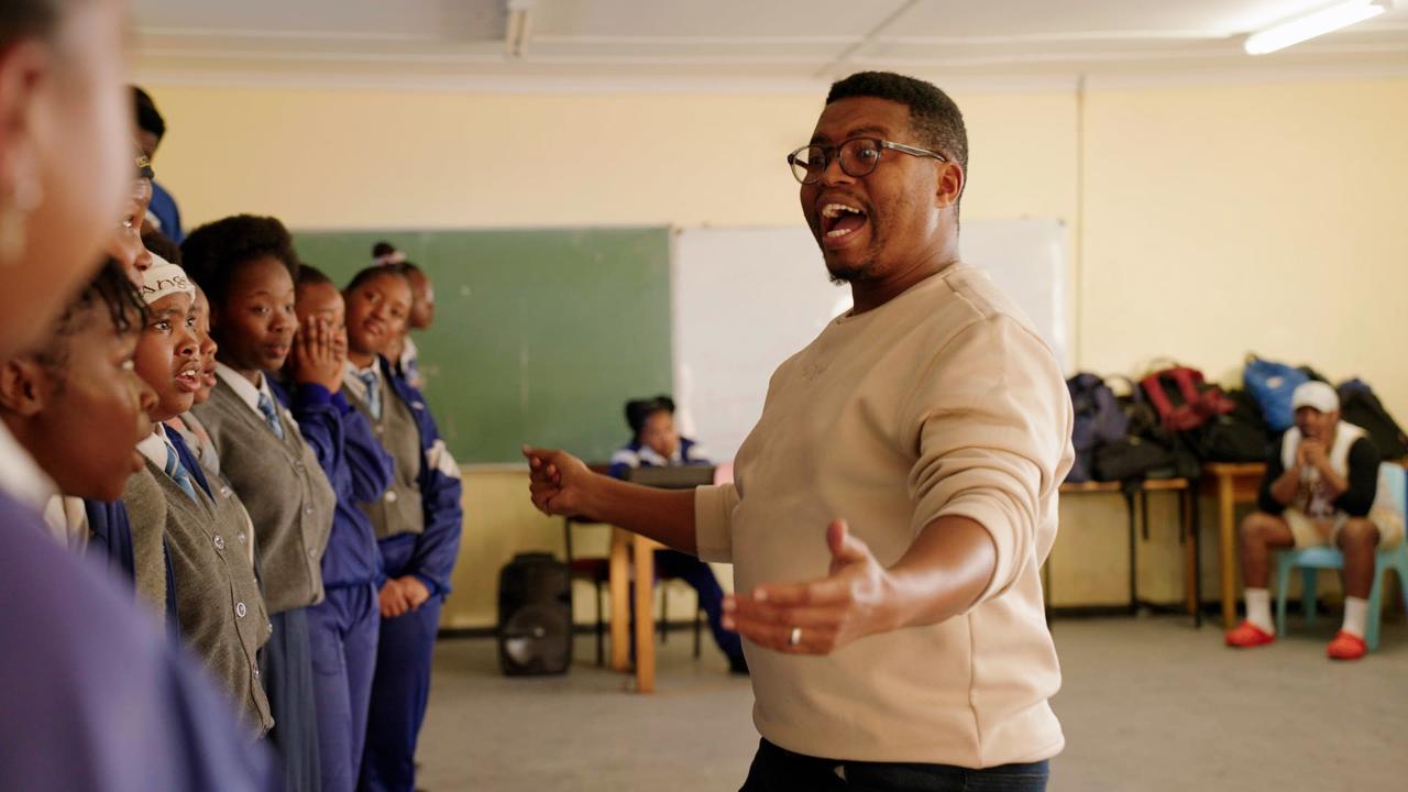 A teacher enthusiastically leading a group of schoolchildren in a classroom, gesturing with open arms while students in uniform stand together and watch him.