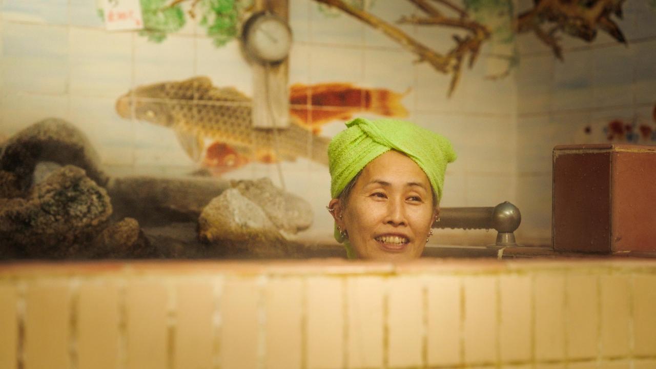 A woman in a green bathing cap sits in a bath with her head appearing over the edge. The side of the bath is tiled with pale yellow sides, with white tiles in the background.