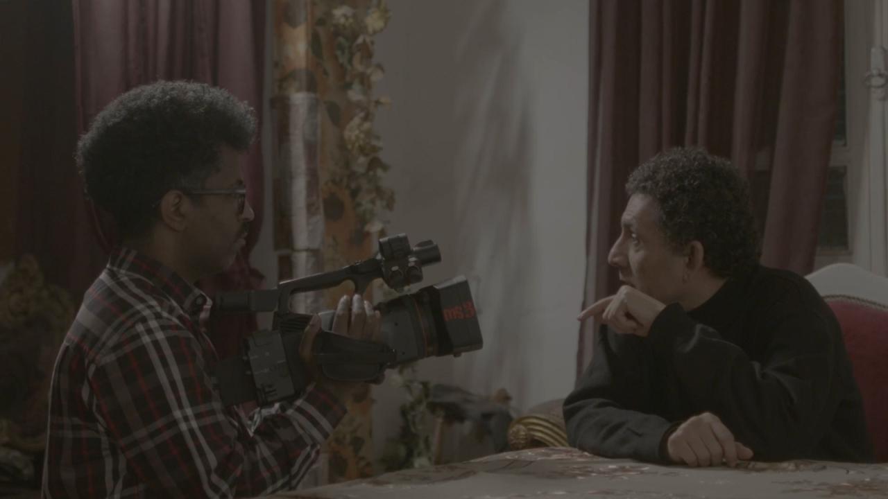 A man in a plaid shirt films another man seated across a table. The second man leans forward thoughtfully, creating an intimate, interview like indoor scene.