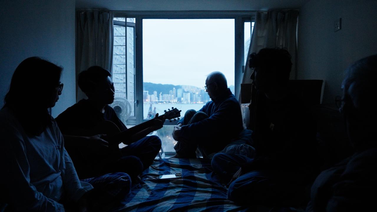 Multiple silhouettes, including one with a guitar centre-left, sit on a bed with a window centrally in the background, framing a view of the sea and city skyline.