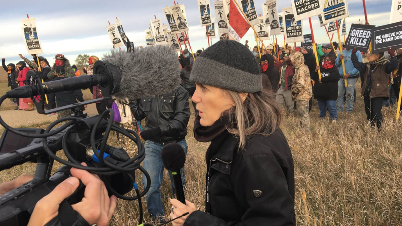 A journalist presents to microphones by a protest.