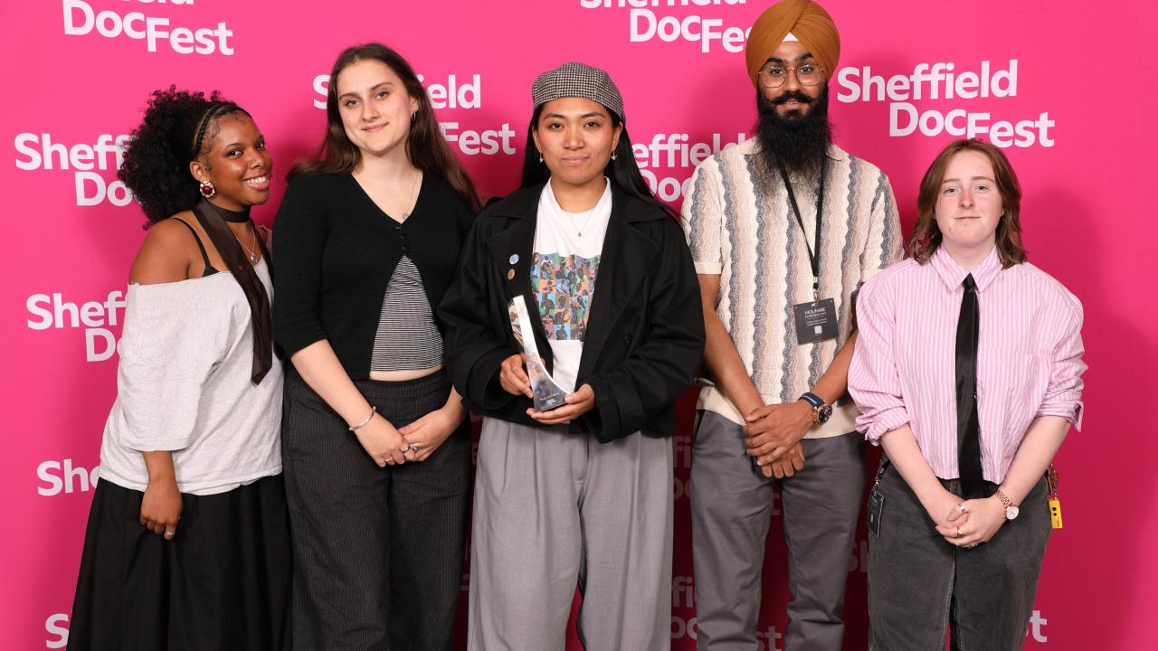 Five young people stand in a line with wide smiles on their faces. Being them is a hot pink step and repeat board with the Sheffield DocFest logo (the words Sheffield DocFest in white) repeatedly pointed on it.