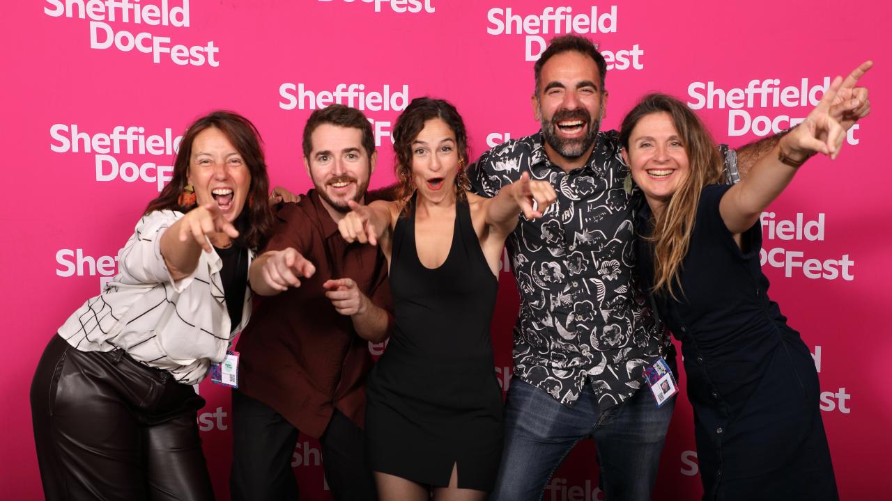Five people point at a camera with wide smiles on their faces.