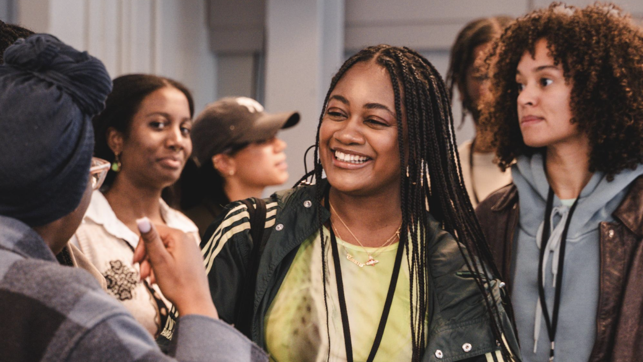  A busy room full of people chatting and smiling. In the centre, a person in a lime green top smiles widely at the person in front of them.