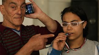 Close-up of a young girl with white glasses biting a medal. The arm of an older man points at the girl as he talks on his mobile phone.